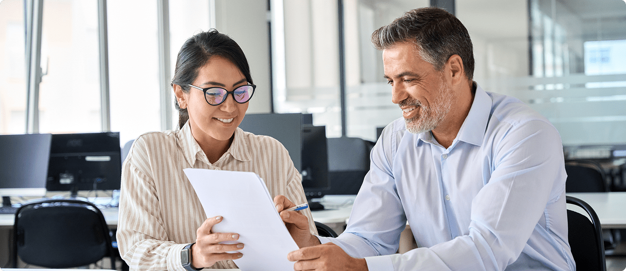 Two people sitting at a desk in an office, looking at documents and discussing paperwork together.